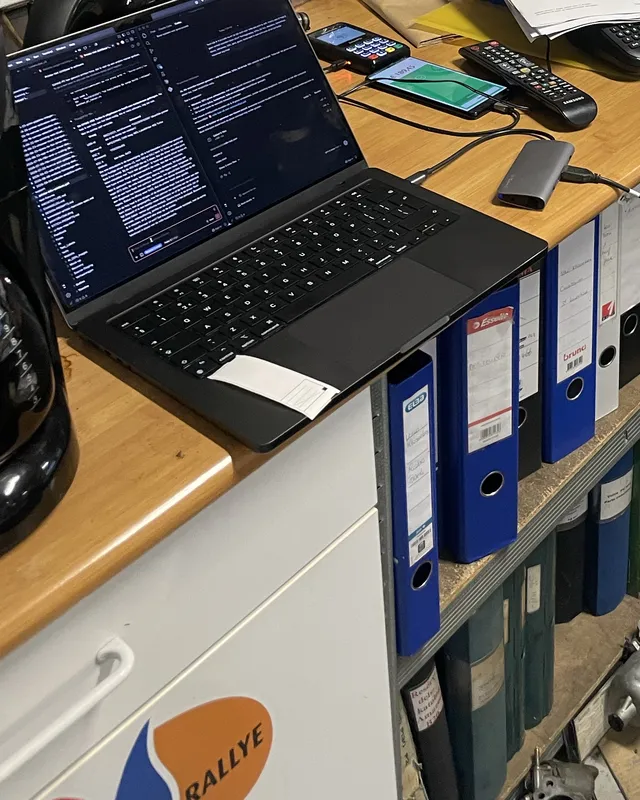 A MacBook on the garage counter with a USB hub and cables, above a row of binders and car parts on the floor