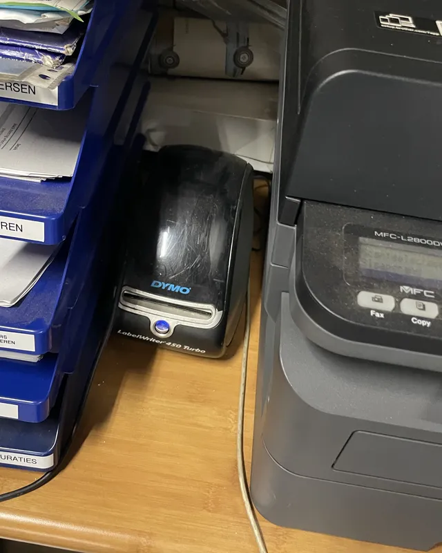 A DYMO LabelWriter 450 Turbo tucked away between blue letter trays full of paper and a large office printer